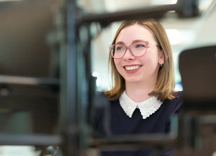 A woman wearing glasses smiling at camera whilst typing at a computer screen in an office setting.